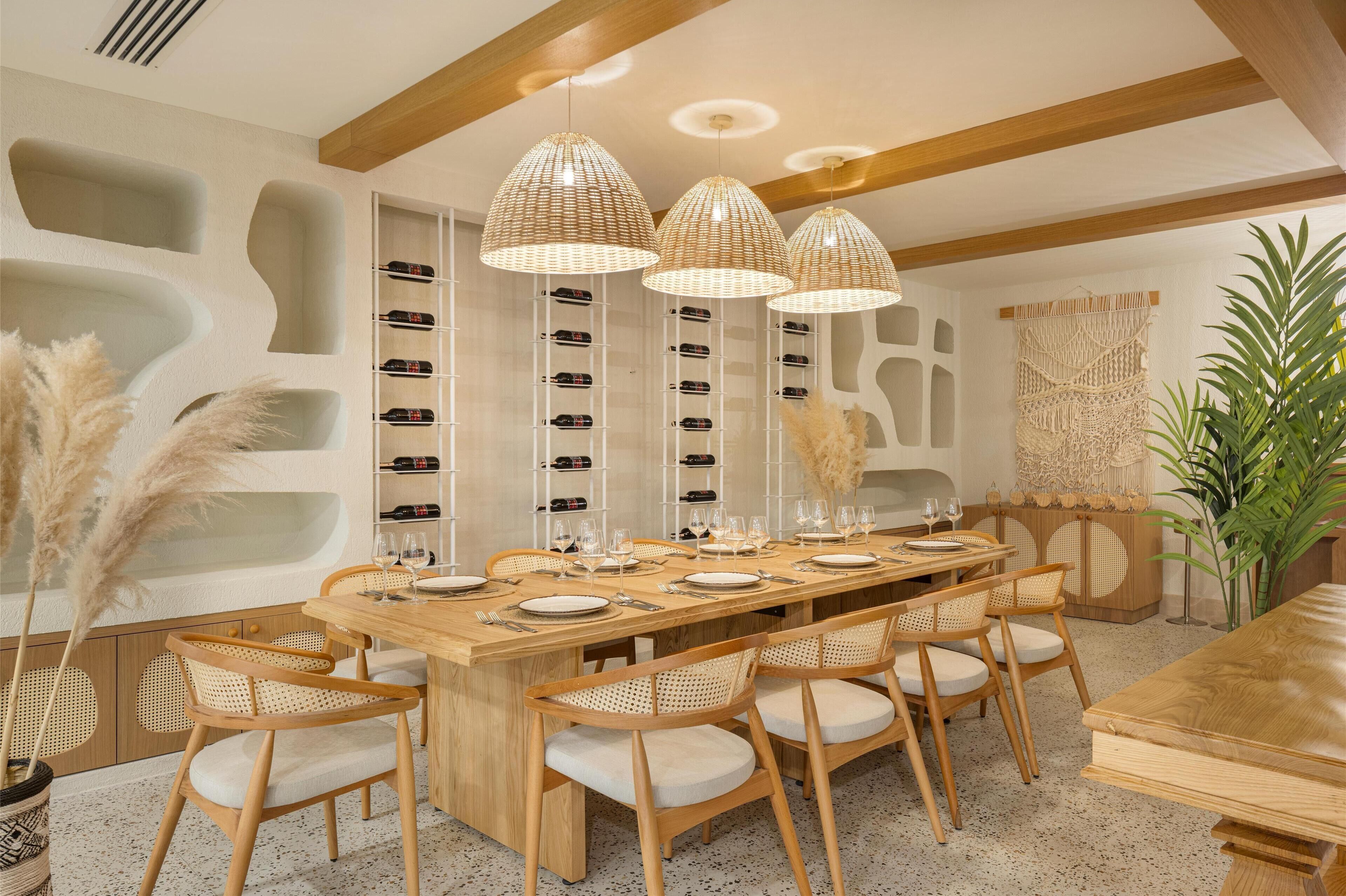 Een eetruimte met een lange houten tafel gedekt voor zes personen, omringd door rieten stoelen met witte kussens. Daarboven hangen drie hanglampen.