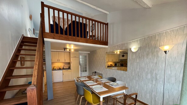 A dining area with a wooden table set for four, a staircase leading to a loft, and a kitchen in the background.