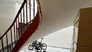 A staircase with a red railing leading to a landing. A bicycle is parked on the landing, and there are boxes stacked against the wall.