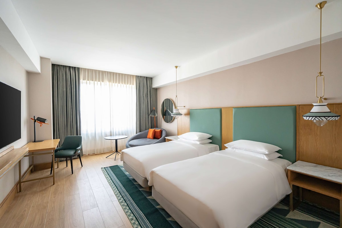 Habitación de hotel con dos camas, un escritorio, una silla, una mesita, un televisor y una ventana con cortinas.