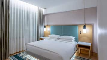 A modern hotel room with a large bed, two bedside tables, a nightstand, a small wooden stool, and a large window with curtains.