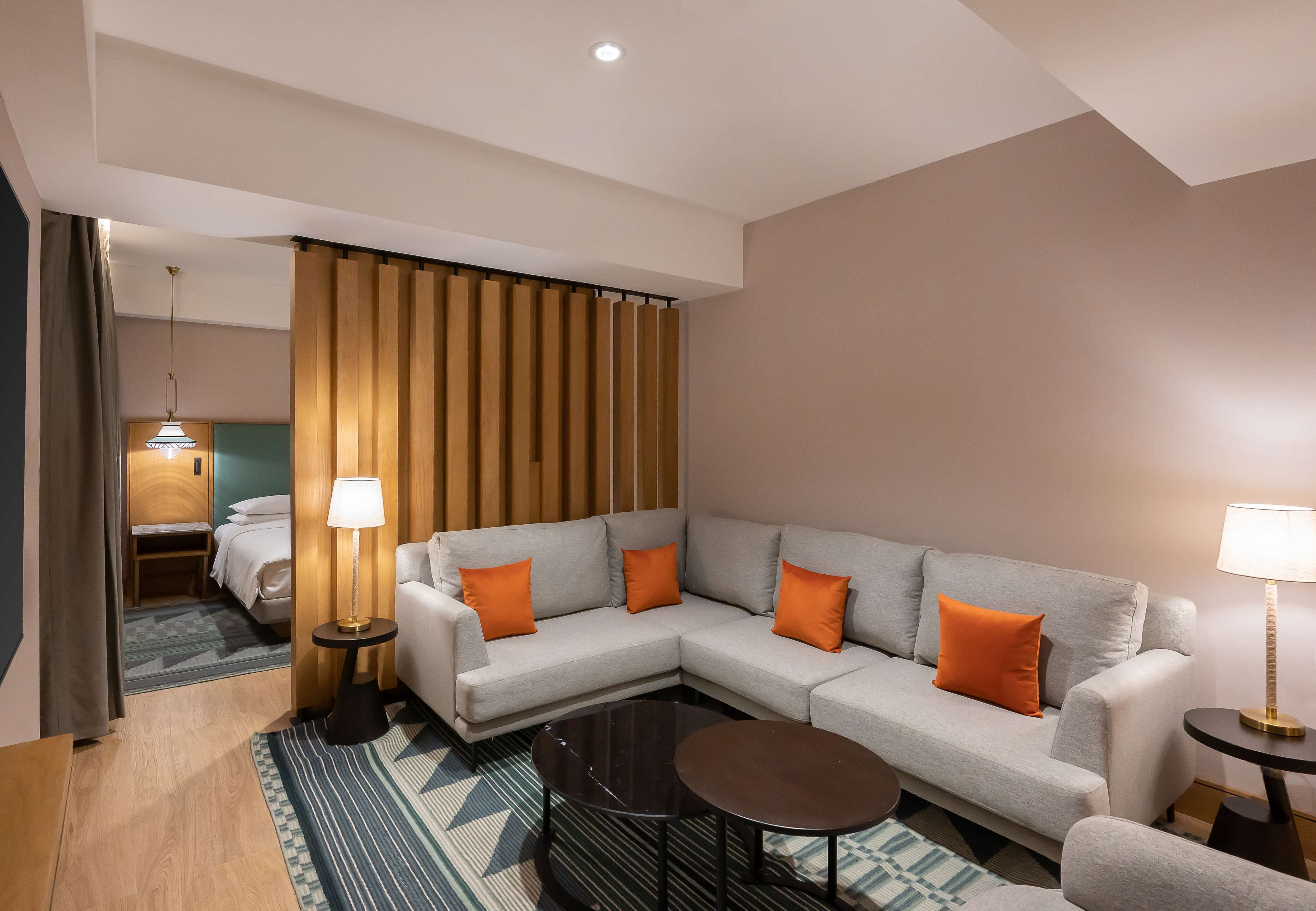 Una habitación de hotel moderna con un sofá seccional, dos mesitas de noche con lámparas, una mesa de centro de cristal, un tapete estampado y un separador de madera.