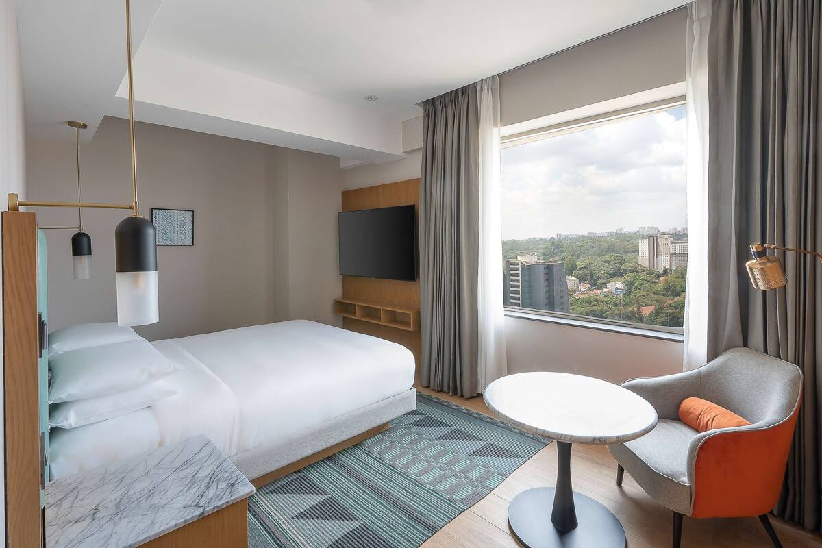 A hotel room with a bed, a TV, a round table, a chair, and a large window with a city view.