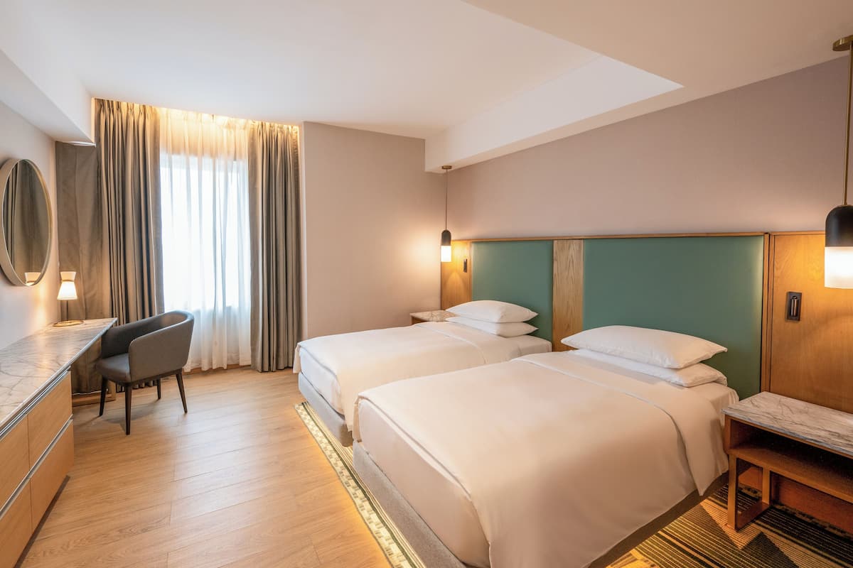 A hotel room with two beds, a desk, a chair, a large window with curtains, and a mirror.