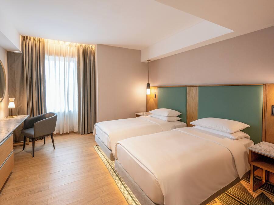 A hotel room with two beds, a desk, a chair, a large window with curtains, and a mirror.