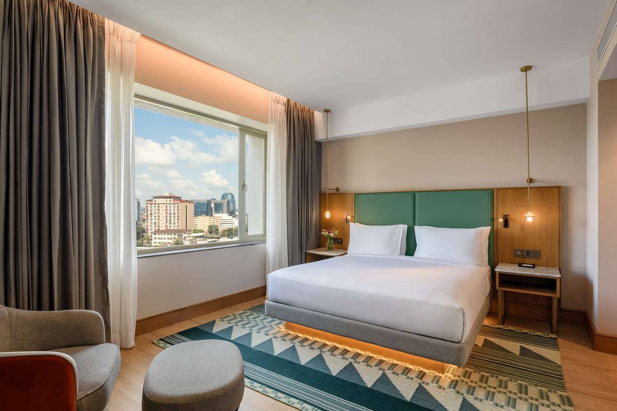 A hotel room with a large bed, a view of the city, a green headboard, and a small table with a lamp.