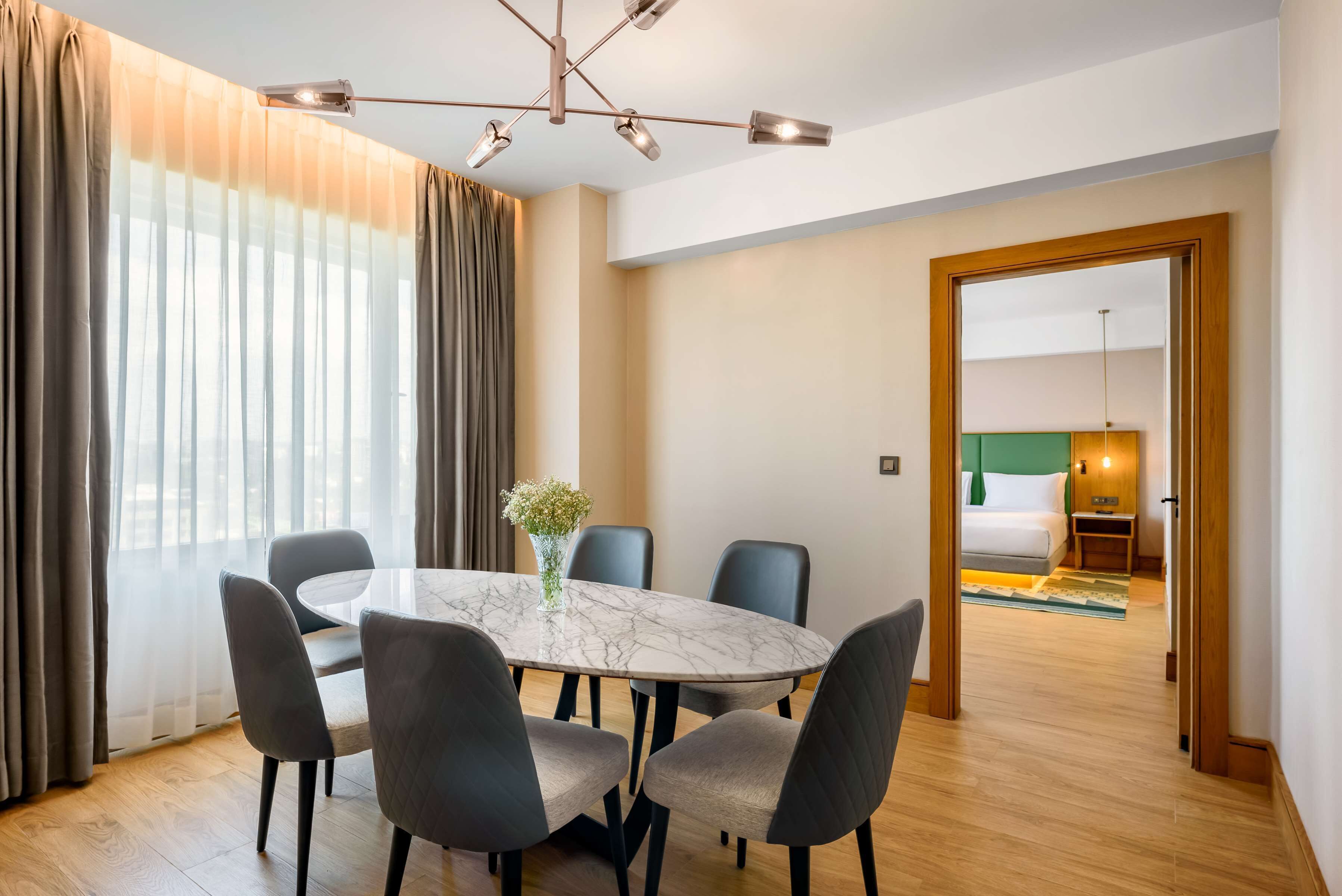 Une chambre d’hôtel dotée d’un coin repas comprenant une table en marbre, des chaises à revêtement gris et un lustre moderne.