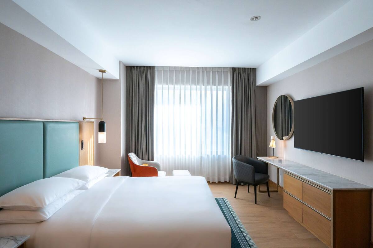 A hotel room with a large bed, a desk, a chair, a TV, and a window with curtains.