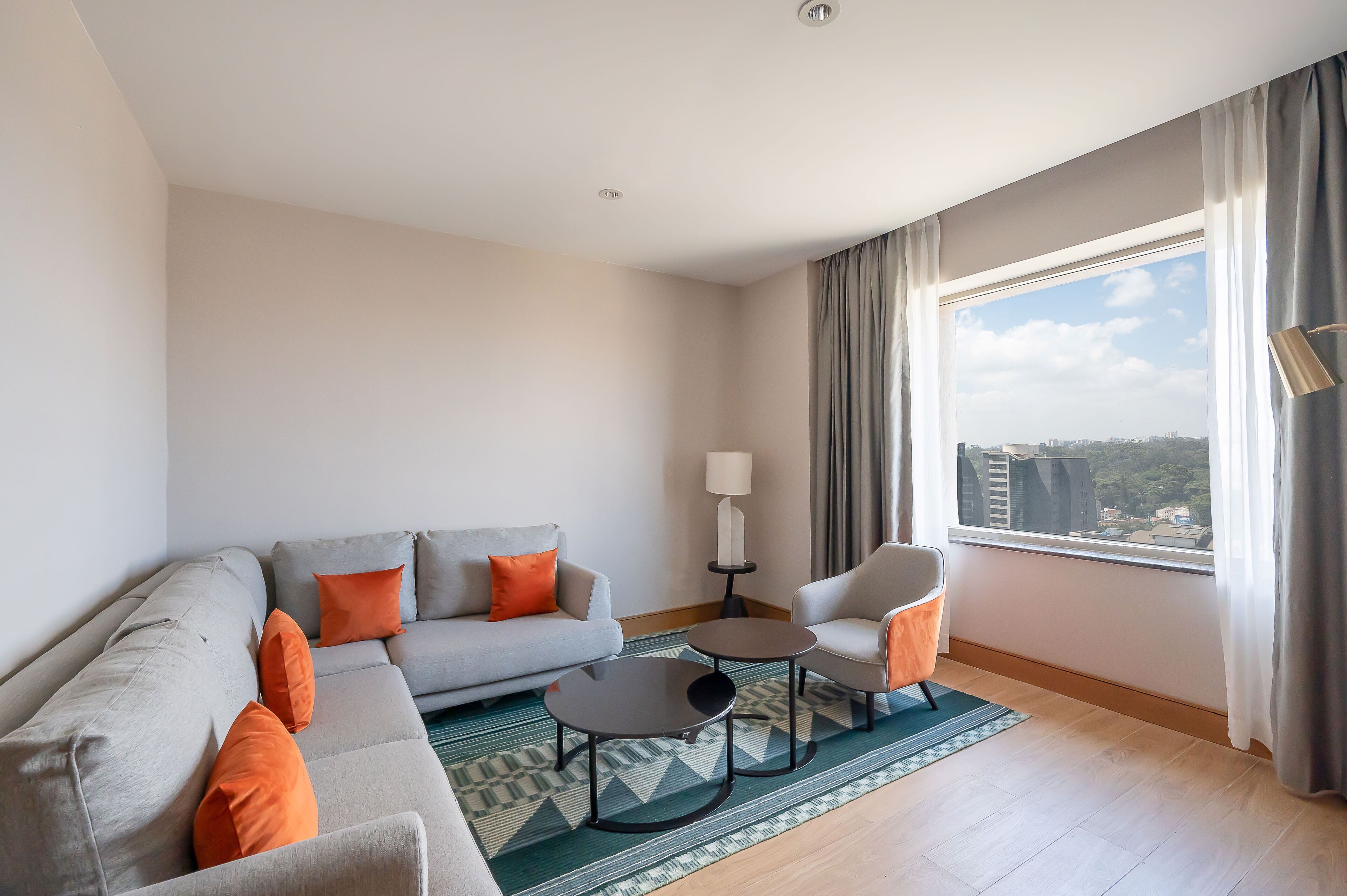 Een moderne woonkamer met een grijze bank, oranje kussens, een klein rond tafeltje en een stoel. Een lamp staat op een bijzettafeltje, en een raam biedt uitzicht op gebouwen en bomen.