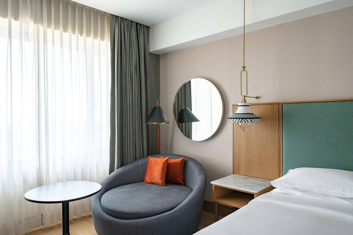 A hotel room with a round mirror, a grey armchair with orange cushions, a small round table, and a bed with white linens.