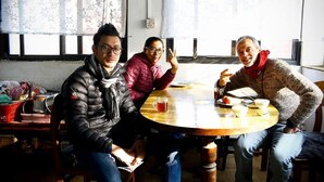 Tres personas sentadas alrededor de una mesa redonda de madera, con una taza de té, en una habitación con cortinas florales y una ventana.