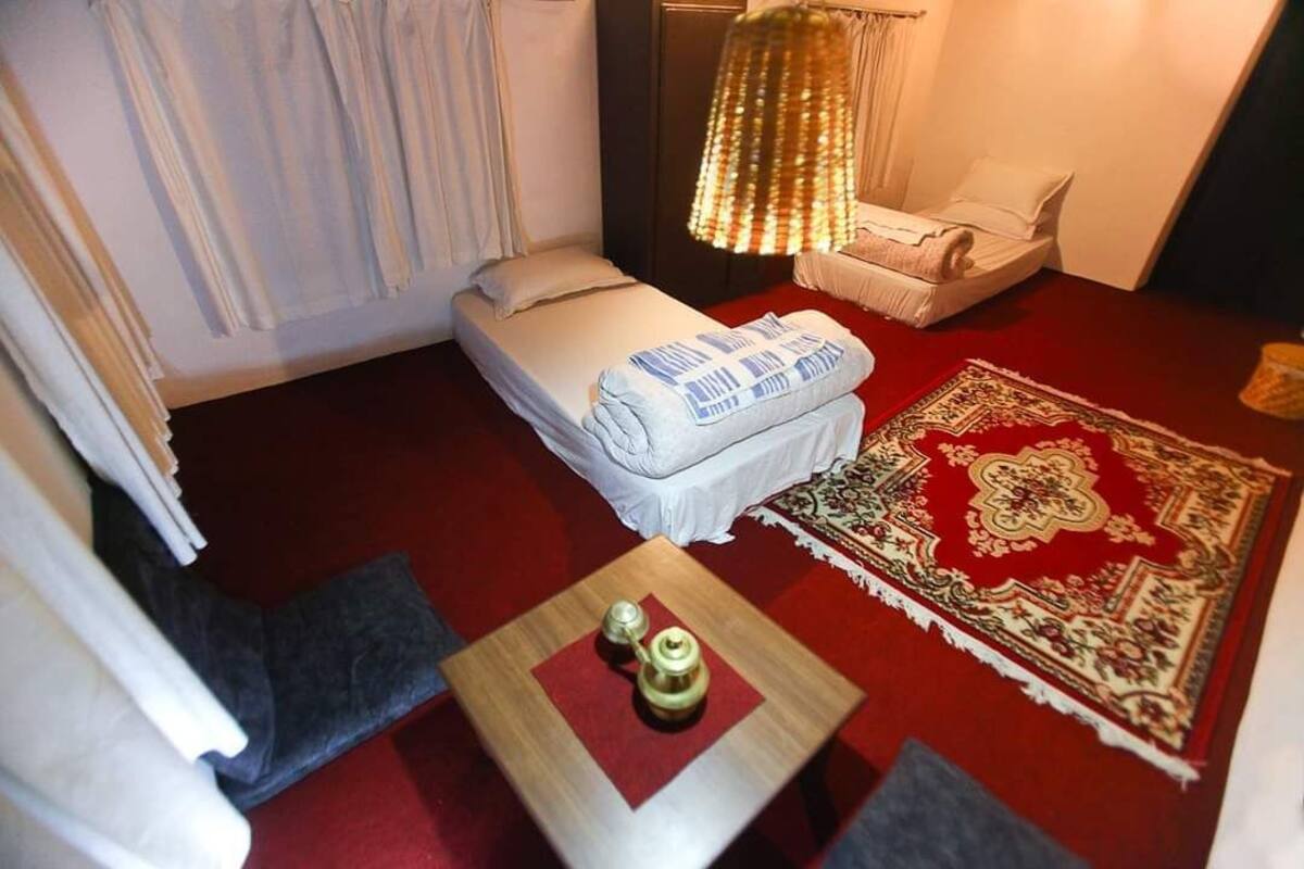 A small, cozy room with a single bed, a small table with a teapot, a red carpet, and a decorative rug.