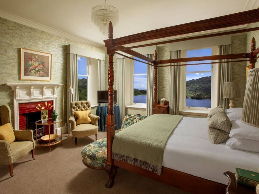 A four-poster bed with a view of a lake and mountains, a fireplace, and a comfortable seating area with a chair and a small table.