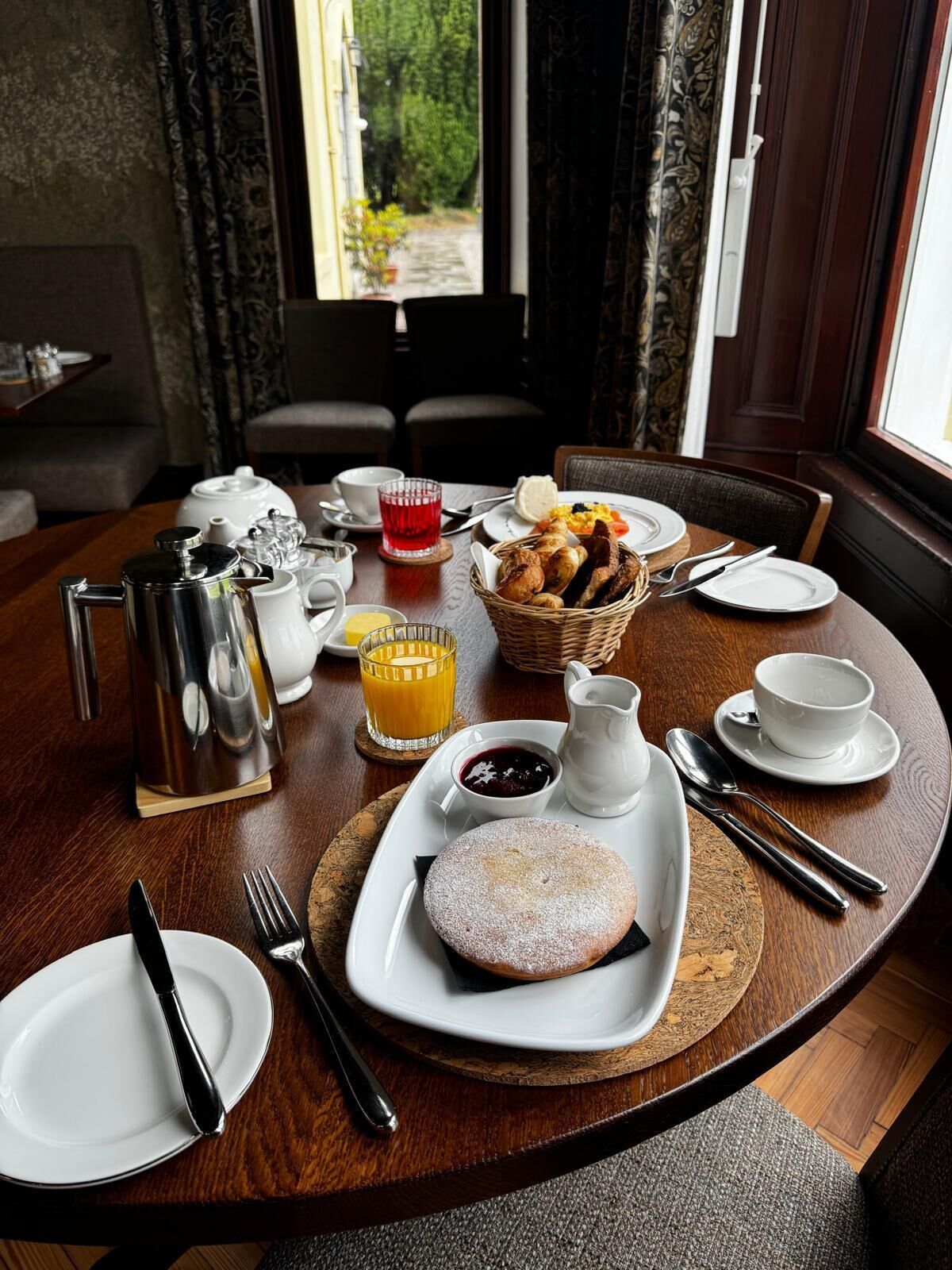 Un desayuno servido con una mesa para dos, incluyendo platos, cubiertos, una tetera, una jarra, una cesta de pan y un postre.