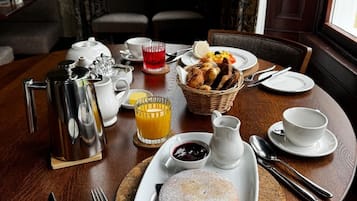 Un desayuno servido con una mesa para dos, incluyendo platos, cubiertos, una tetera, una jarra, una cesta de pan y un postre.