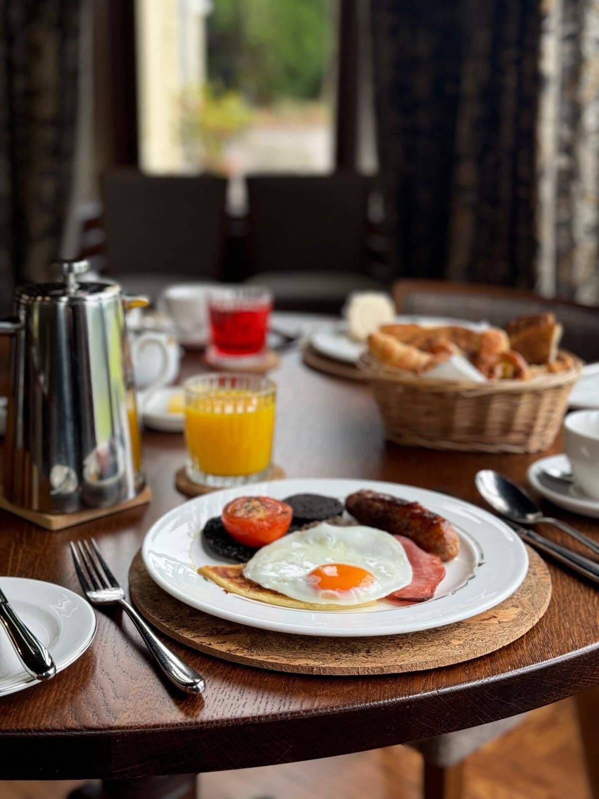 A breakfast setting with a plate of eggs, sausages, and toast, a coffee pot, a glass of orange juice, and a basket of bread on a wooden table.