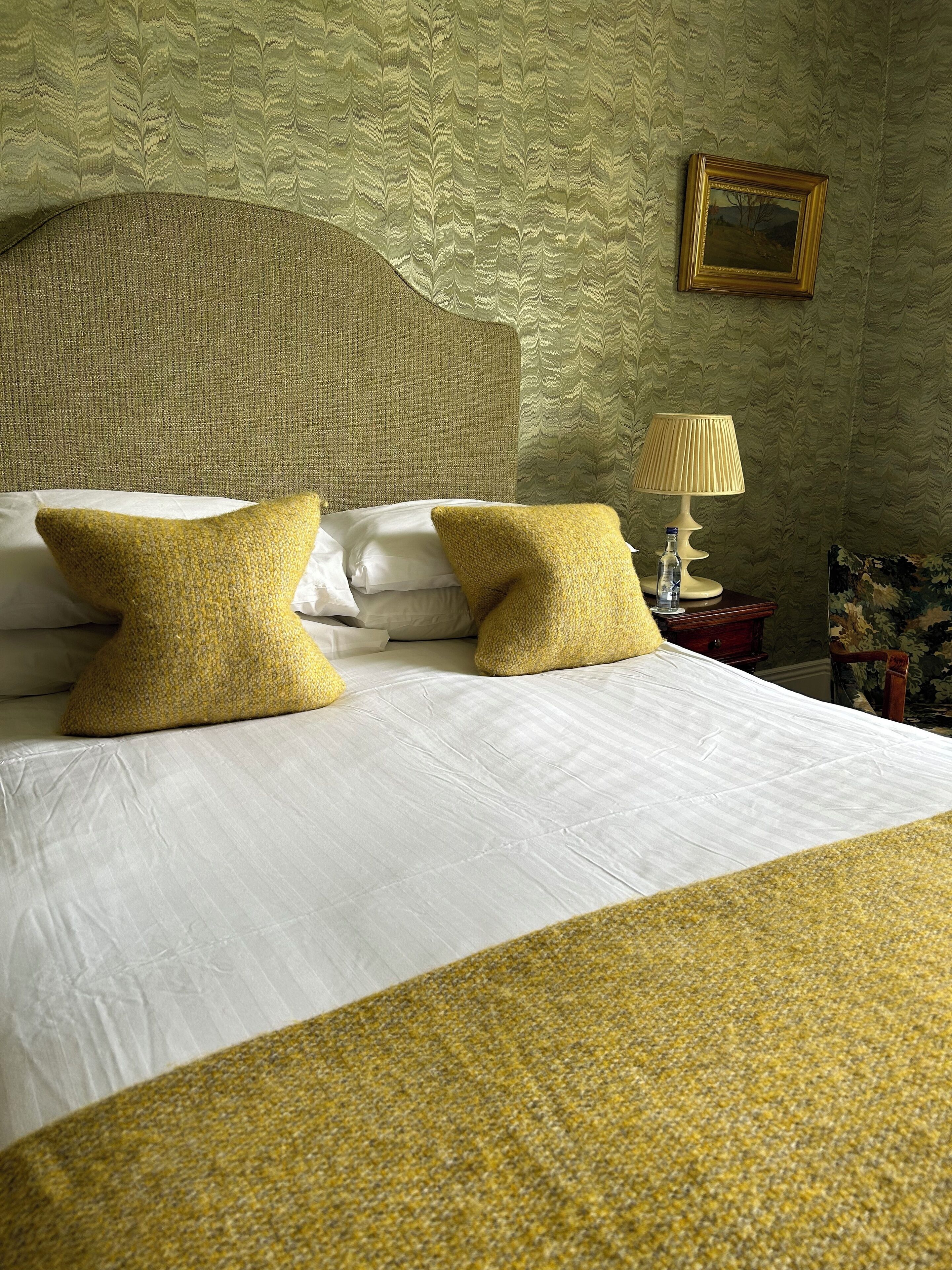 A neatly made bed with white linens and two yellow pillows, a wooden headboard, a bedside table with a lamp and a bottle, and a framed picture on the wall.
