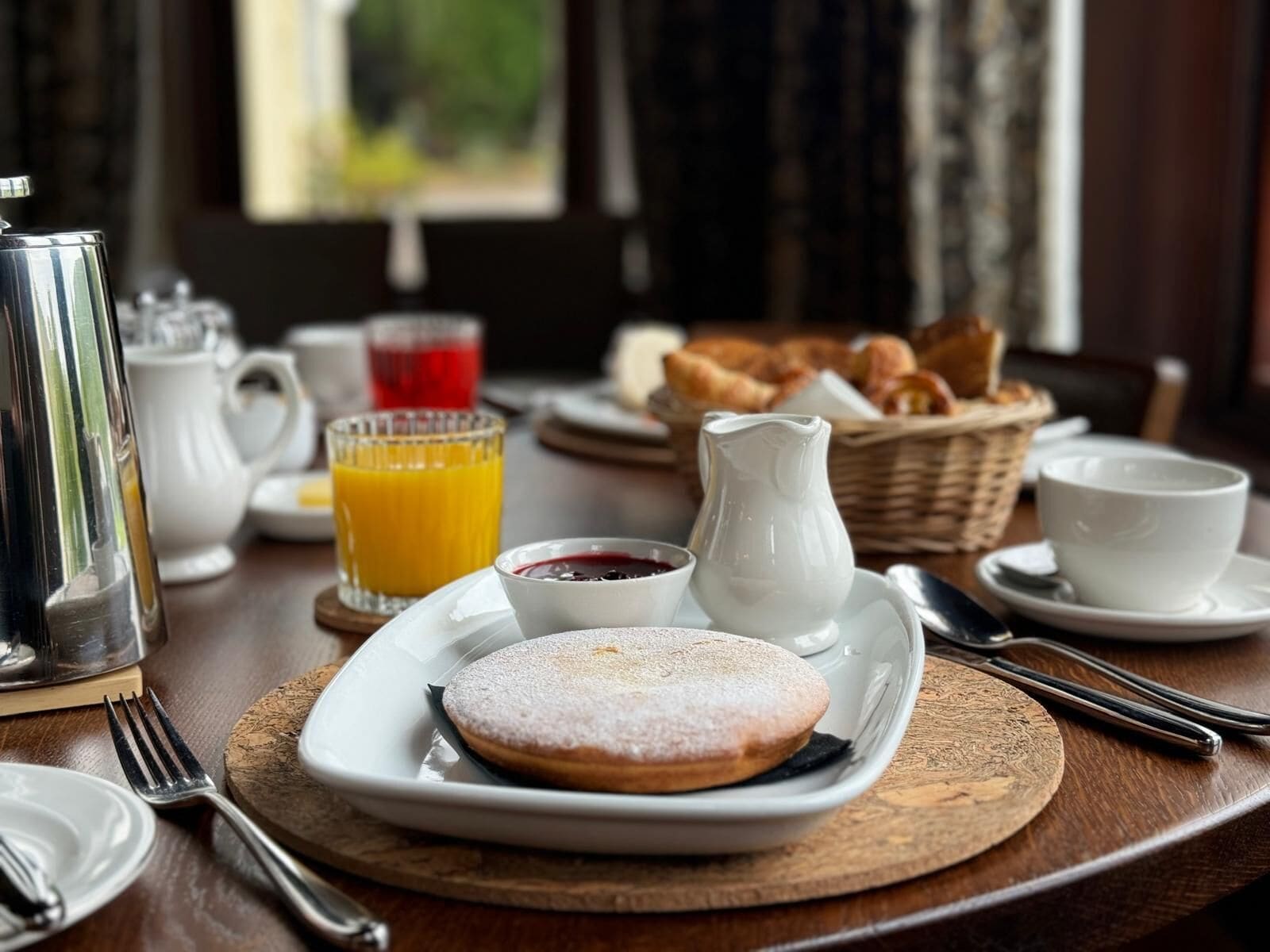 Una mesa de desayuno con un plato de pasteles, una jarra de crema, un vaso de zumo de naranja y una cesta de croissants.