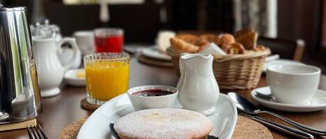 Una mesa de desayuno con un plato de pasteles, una jarra de crema, un vaso de zumo de naranja y una cesta de croissants.