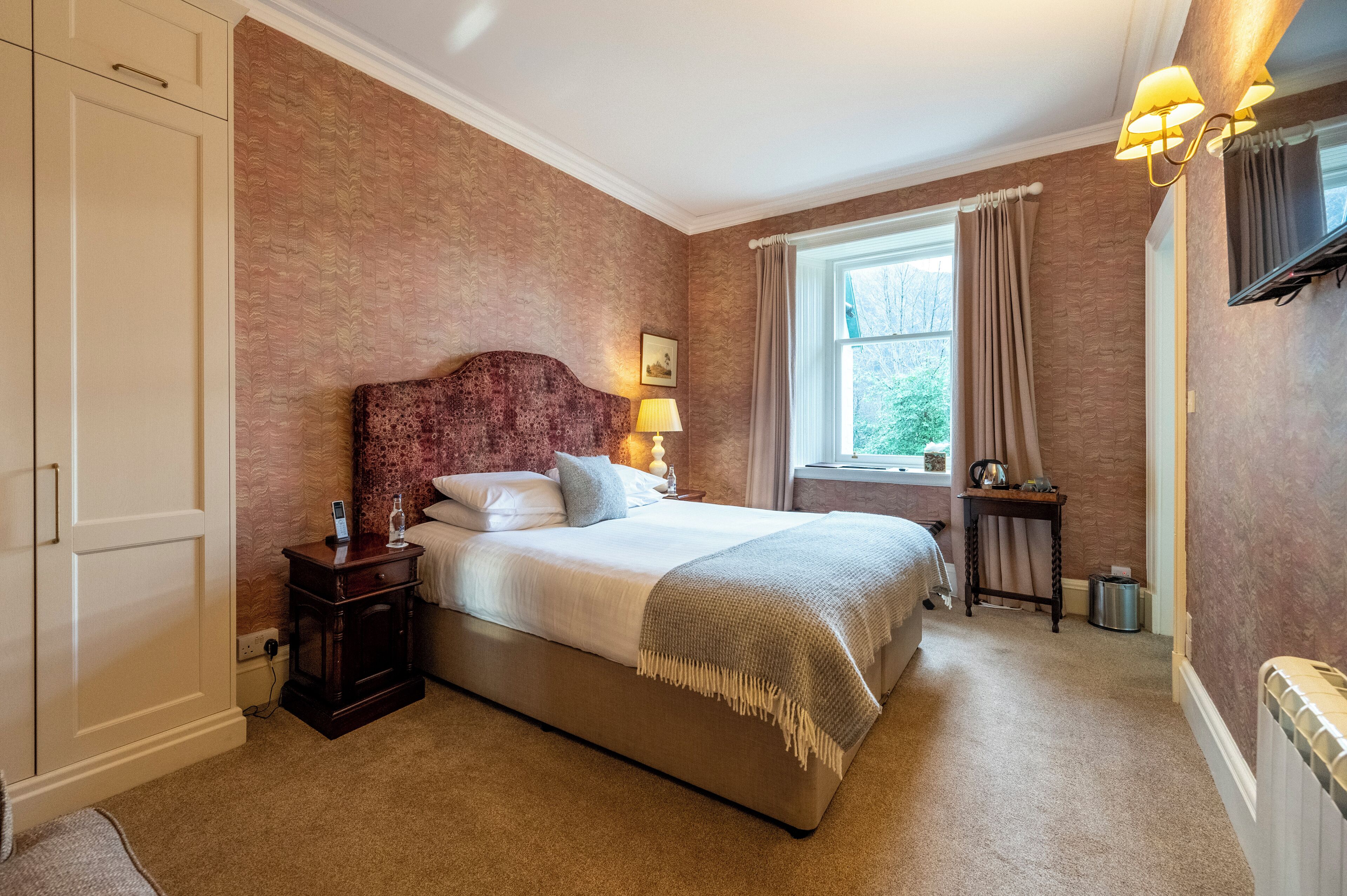 A bedroom with a large bed, a nightstand, a window with curtains, and a radiator.