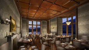 Ein Restaurant mit Holzbalkendecke, großen Fenstern und Blick auf Berge. Die Tische sind mit Tellern, Gläsern und Besteck gedeckt.