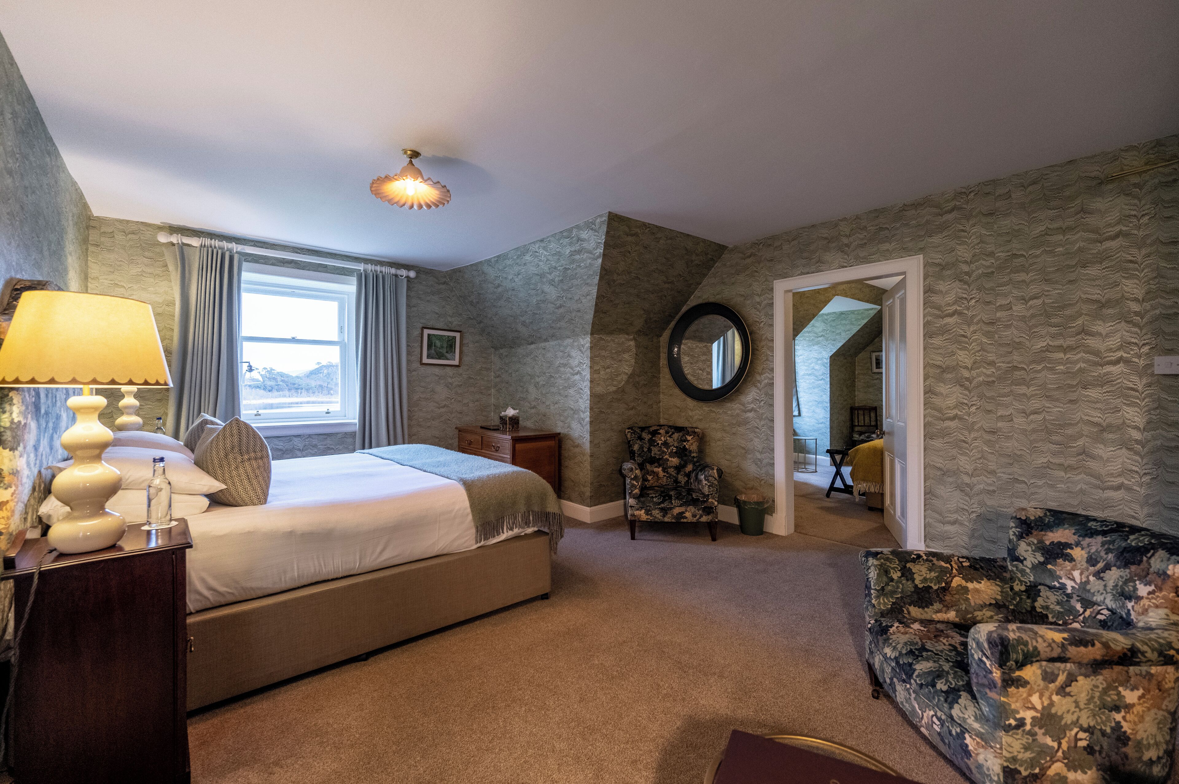 A hotel room with a large bed, a chair, a lamp, and a view of the mountains through the window.