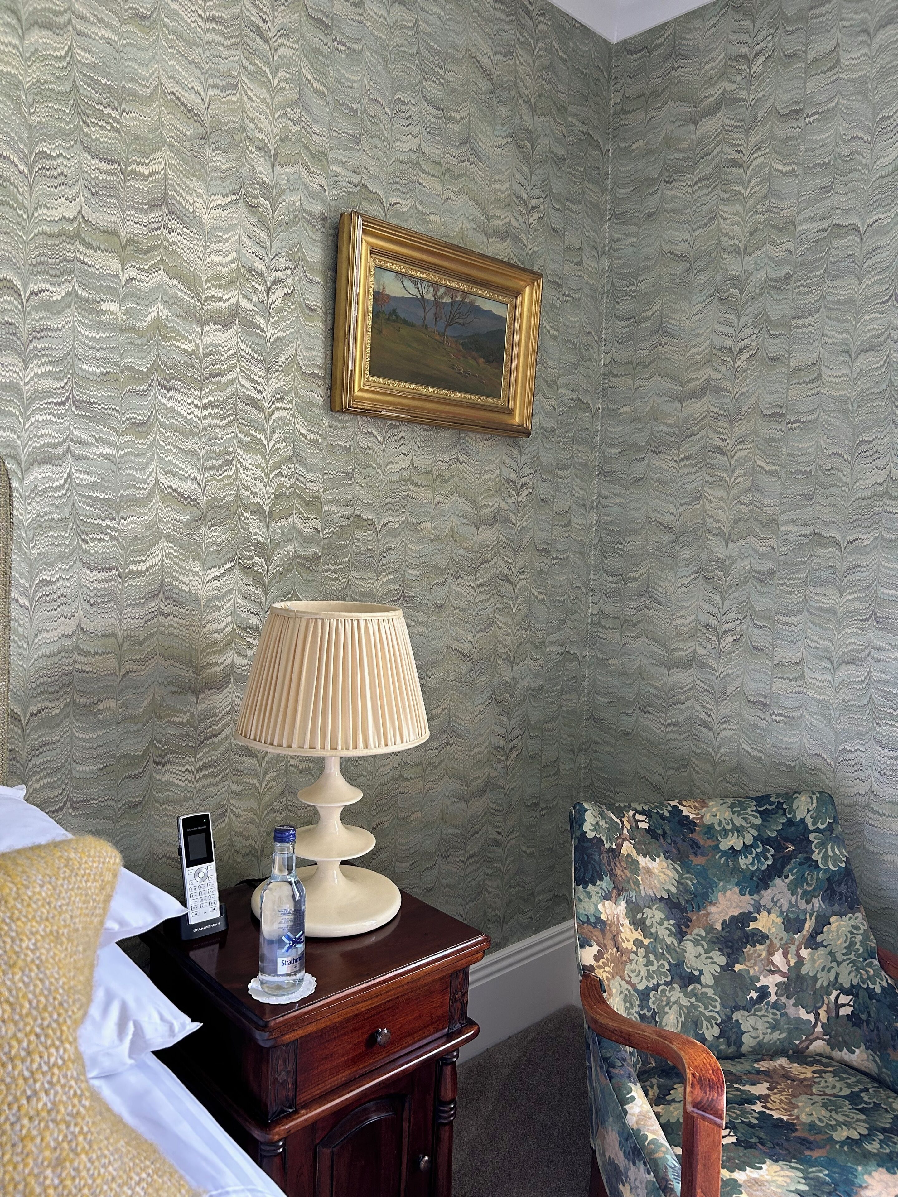 A hotel room with a patterned wallpaper, a bedside table with a lamp, a chair, and a framed picture on the wall.