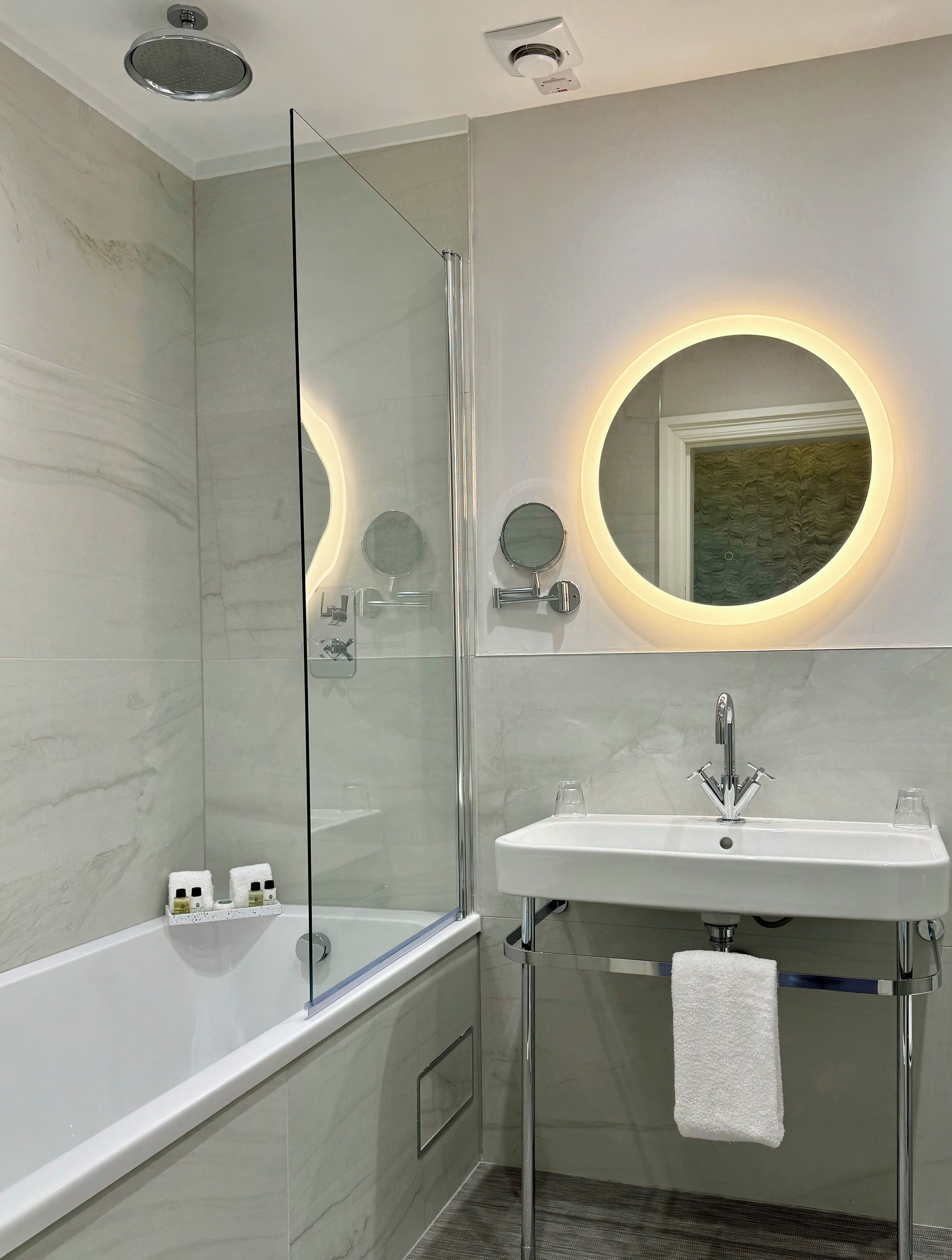 A modern bathroom with a glass-enclosed shower, a white sink with a towel, and a round mirror with a light.