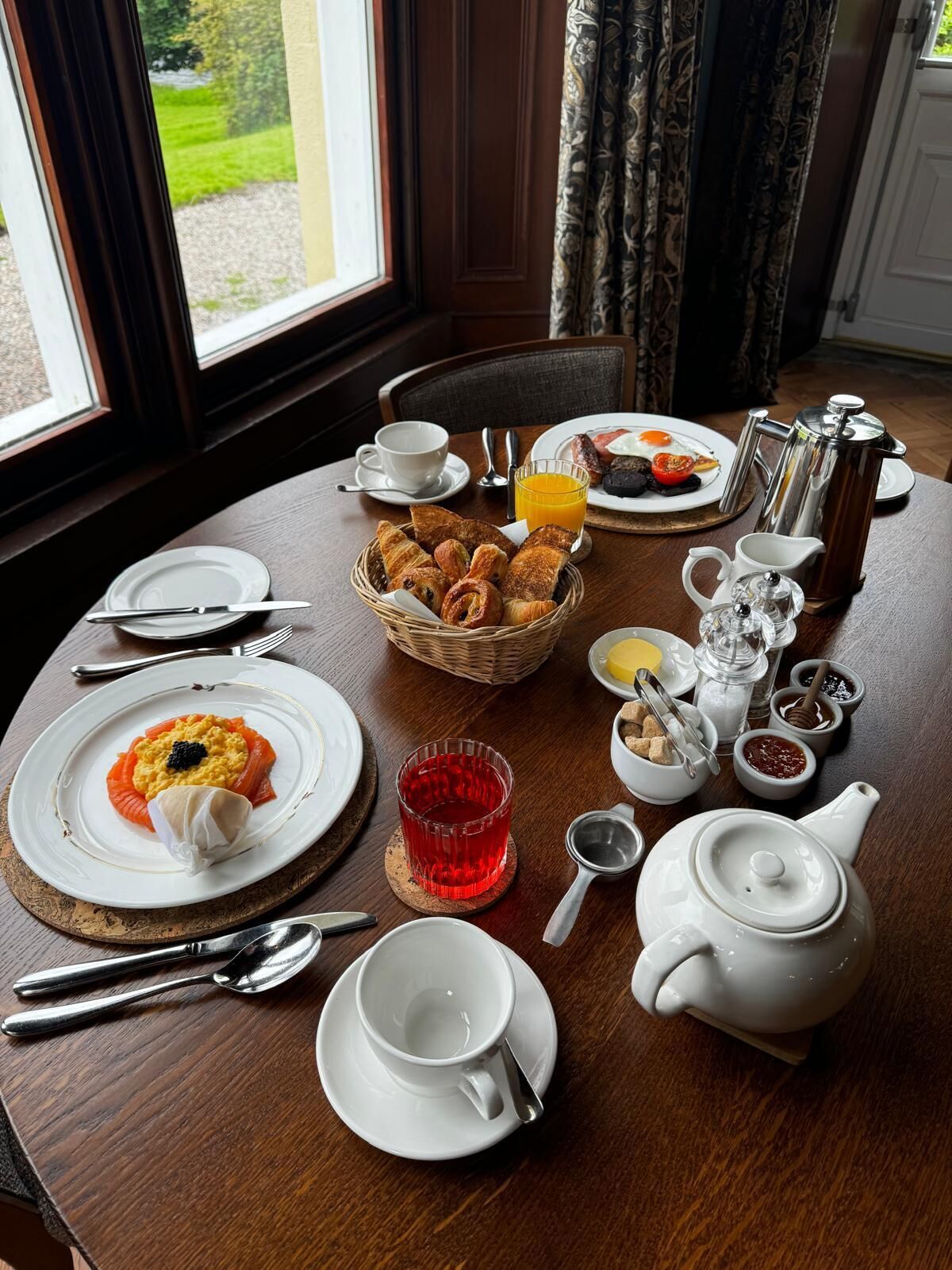 A breakfast table with plates of food, a coffee pot, a teapot, and a glass of orange juice.