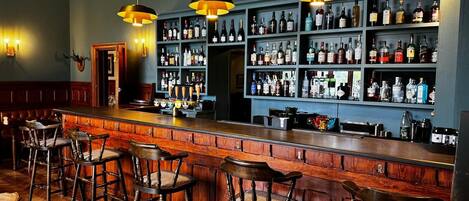 A bar area with wooden bar stools, a counter with a tap, and shelves stocked with bottles.