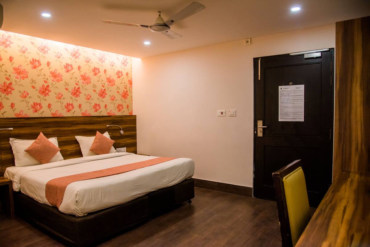 A hotel room with a bed, wooden headboard, floral wallpaper, a ceiling fan, a door with a notice, and a wooden table.