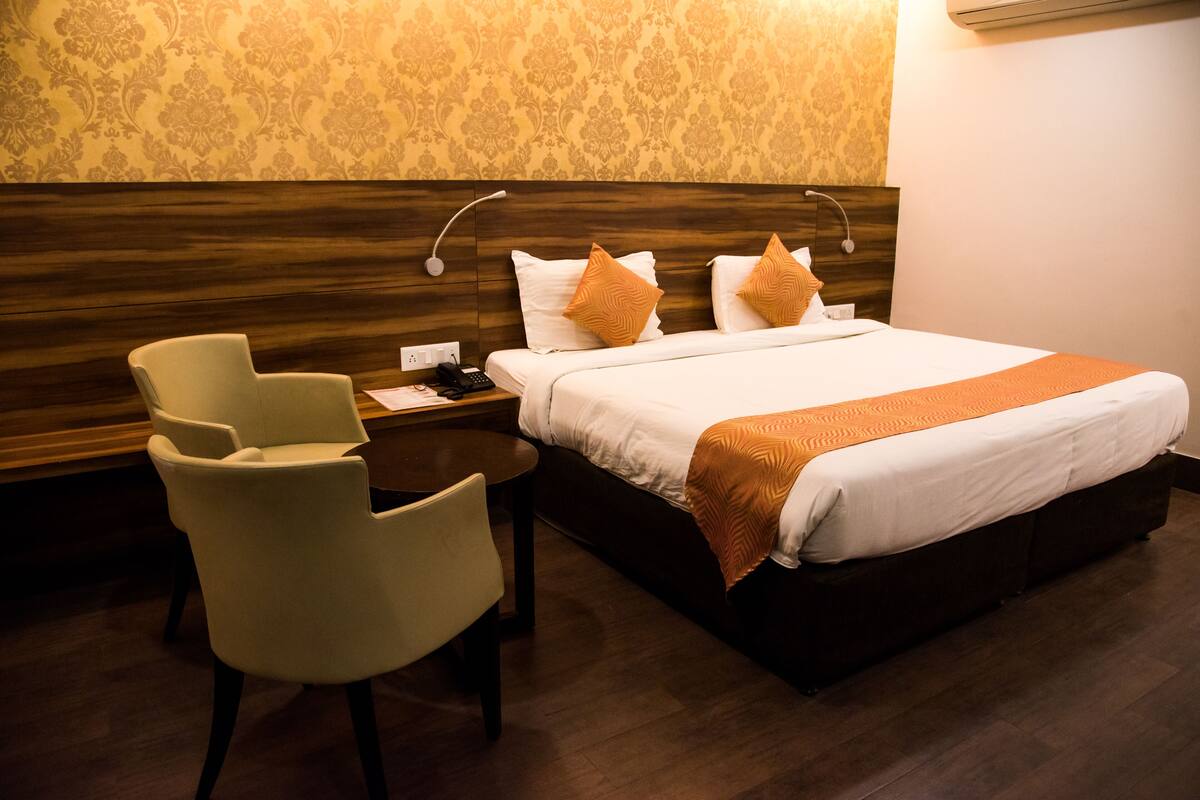 A hotel room with a bed, two pillows, a bedside table with a phone, a chair, and a wall with patterned wallpaper.