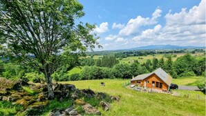 Une cabane en bois, dotée d’un toit à pente, entourée de champs verdoyants et d’arbres, sous un ciel dégagé.
