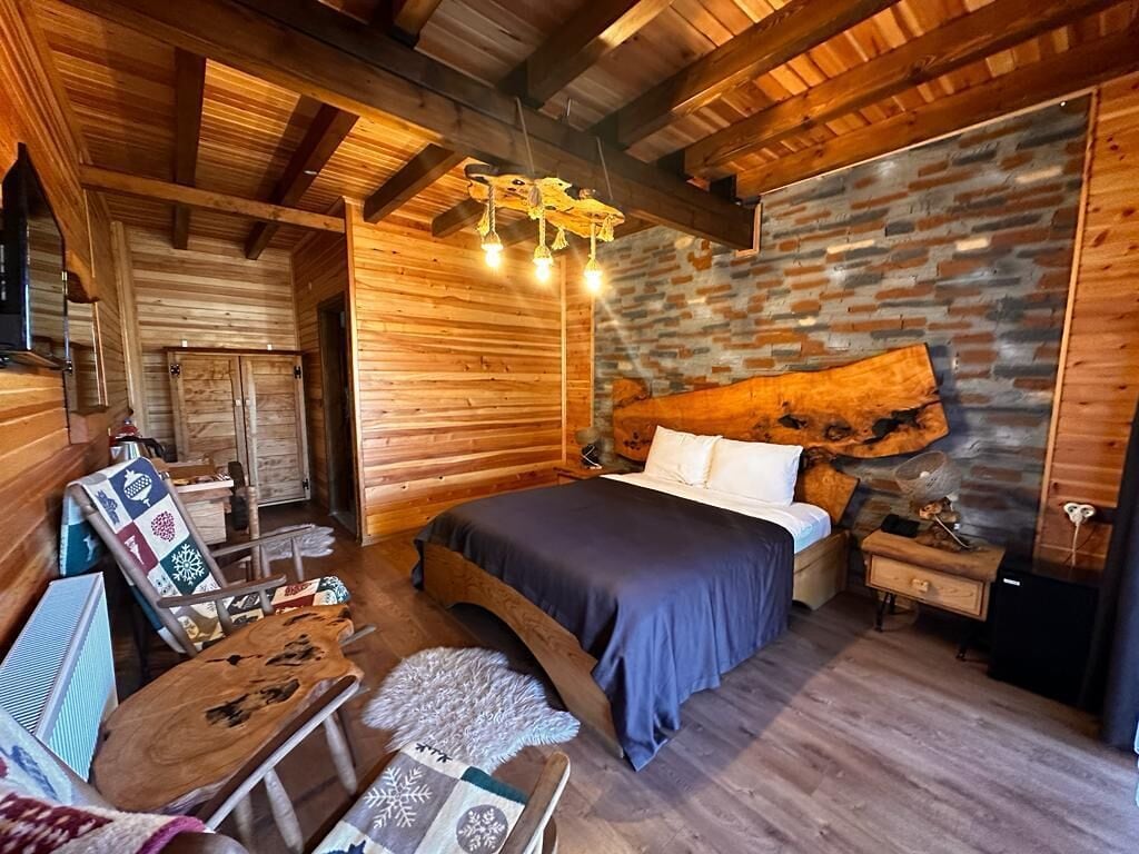 A cozy log cabin room with a bed, wooden walls, a stone accent wall, and a rustic wooden table.