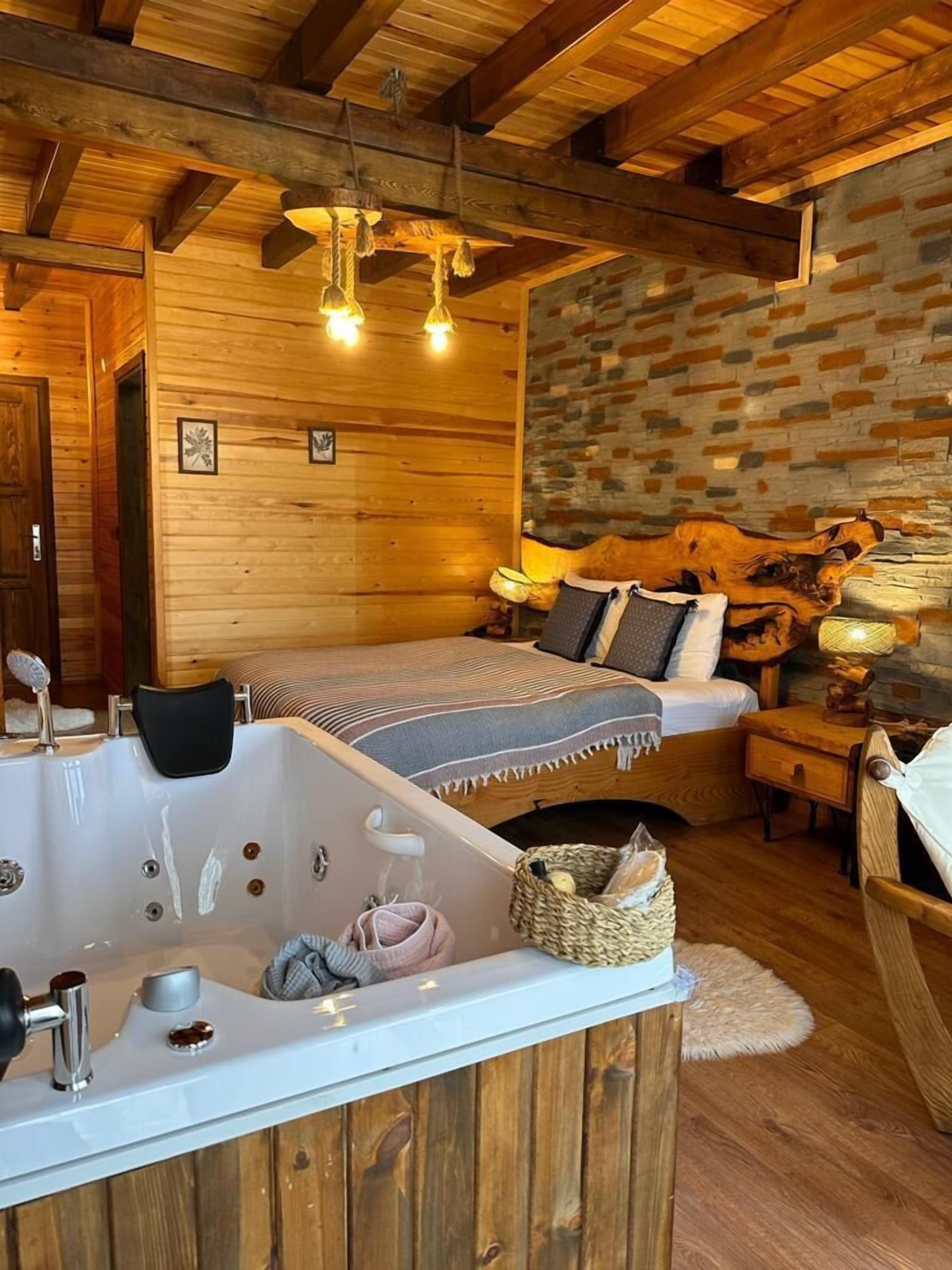 A cozy room with a wooden ceiling, a stone wall, a bed with pillows, a nightstand, a lamp, and a bathtub with towels and a basket.