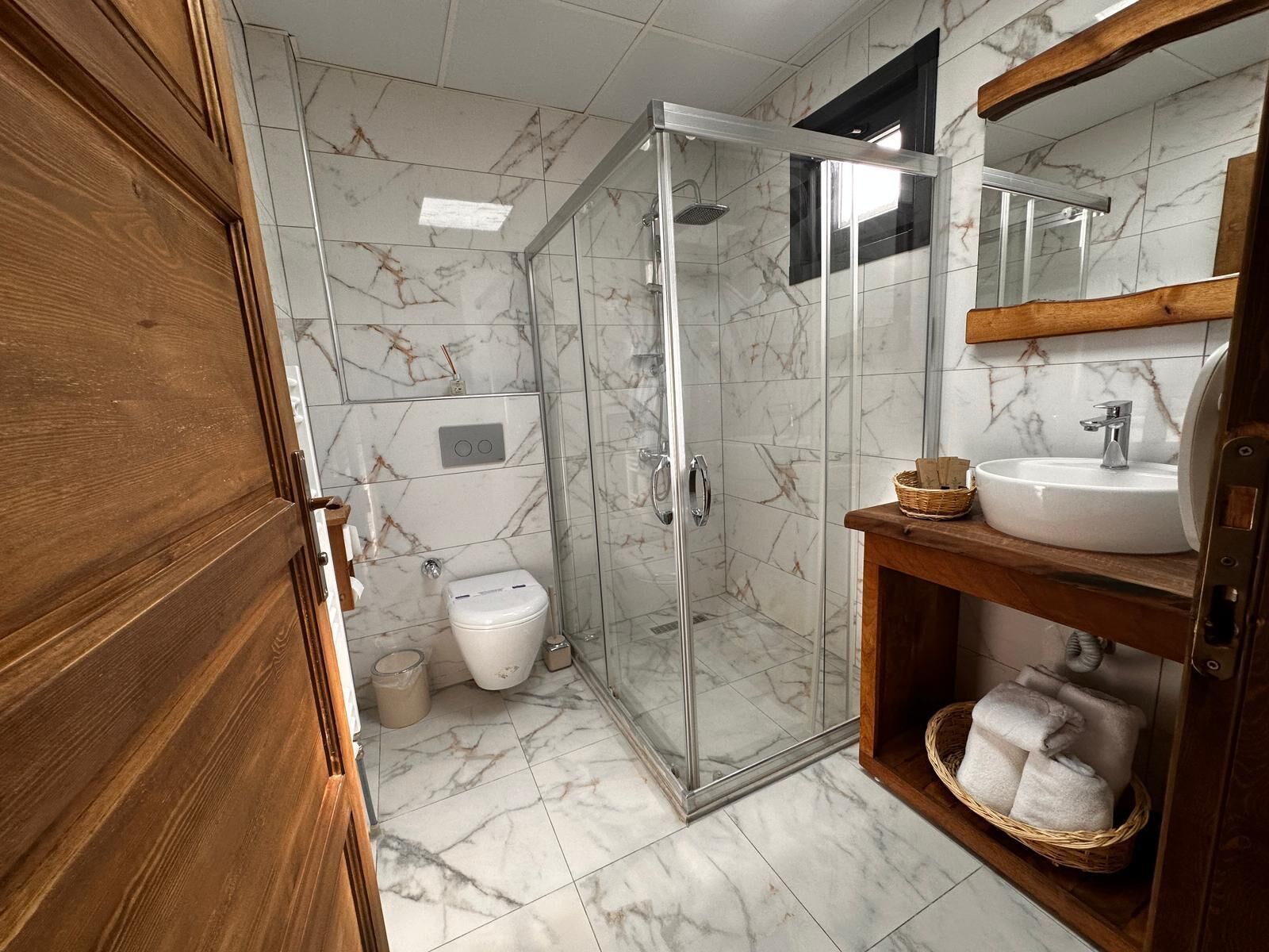A bathroom with a glass-enclosed shower, a white toilet, and a wooden vanity with a sink and a basket of towels.
