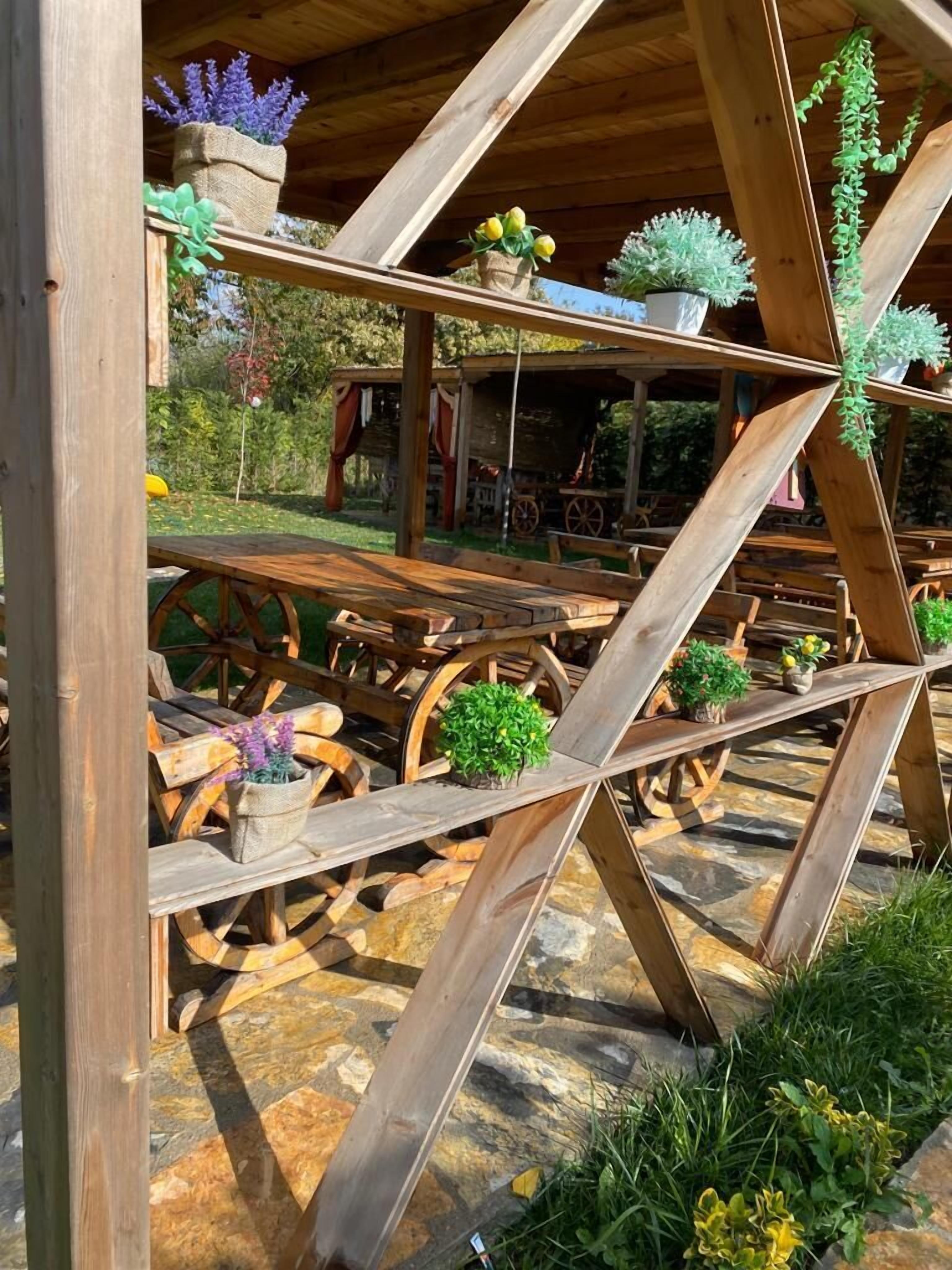 Une structure rustique en bois, dotée d’un sol en pierre, de tables et de chaises en bois, et entourée de plantes en pot et de verdure.