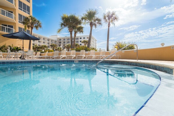 A clear swimming pool with lounge chairs and umbrellas, surrounded by palm trees and a multi-story building.