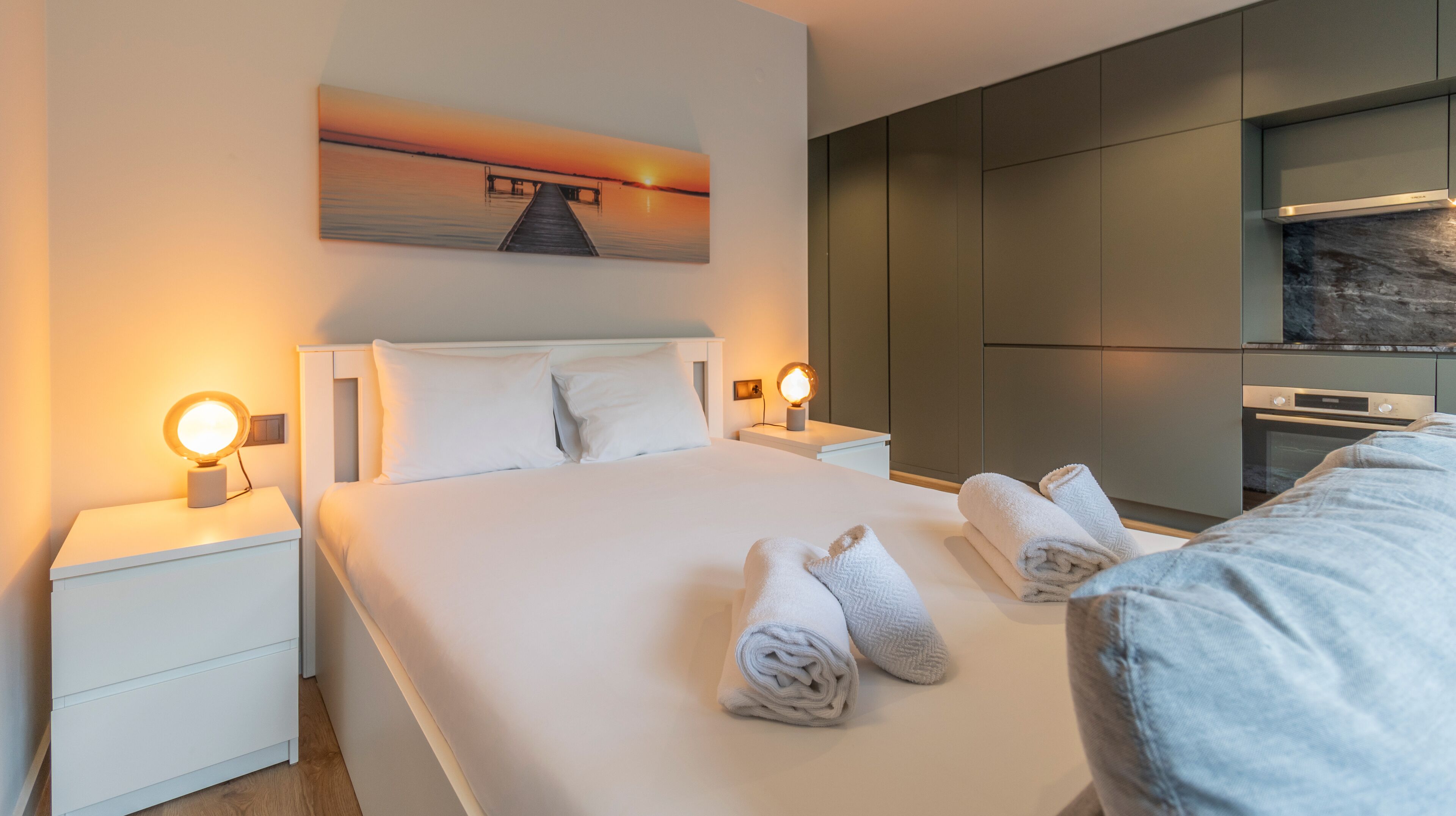 Ein modernes Hotelzimmer mit einem großen Bett, zwei Nachttischlampen, einem Sonnenuntergangsgemälde und einer integrierten Küchenzeile.