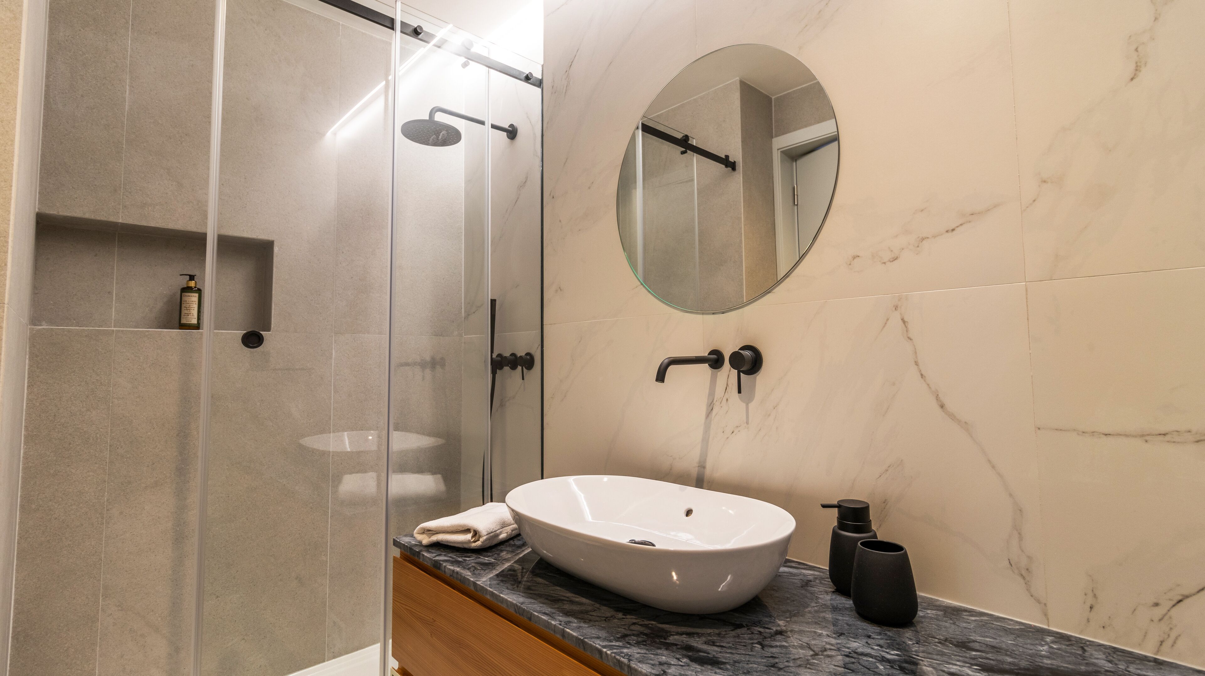 A modern bathroom with a glass-enclosed shower, a round mirror, a white sink, and a marble countertop.