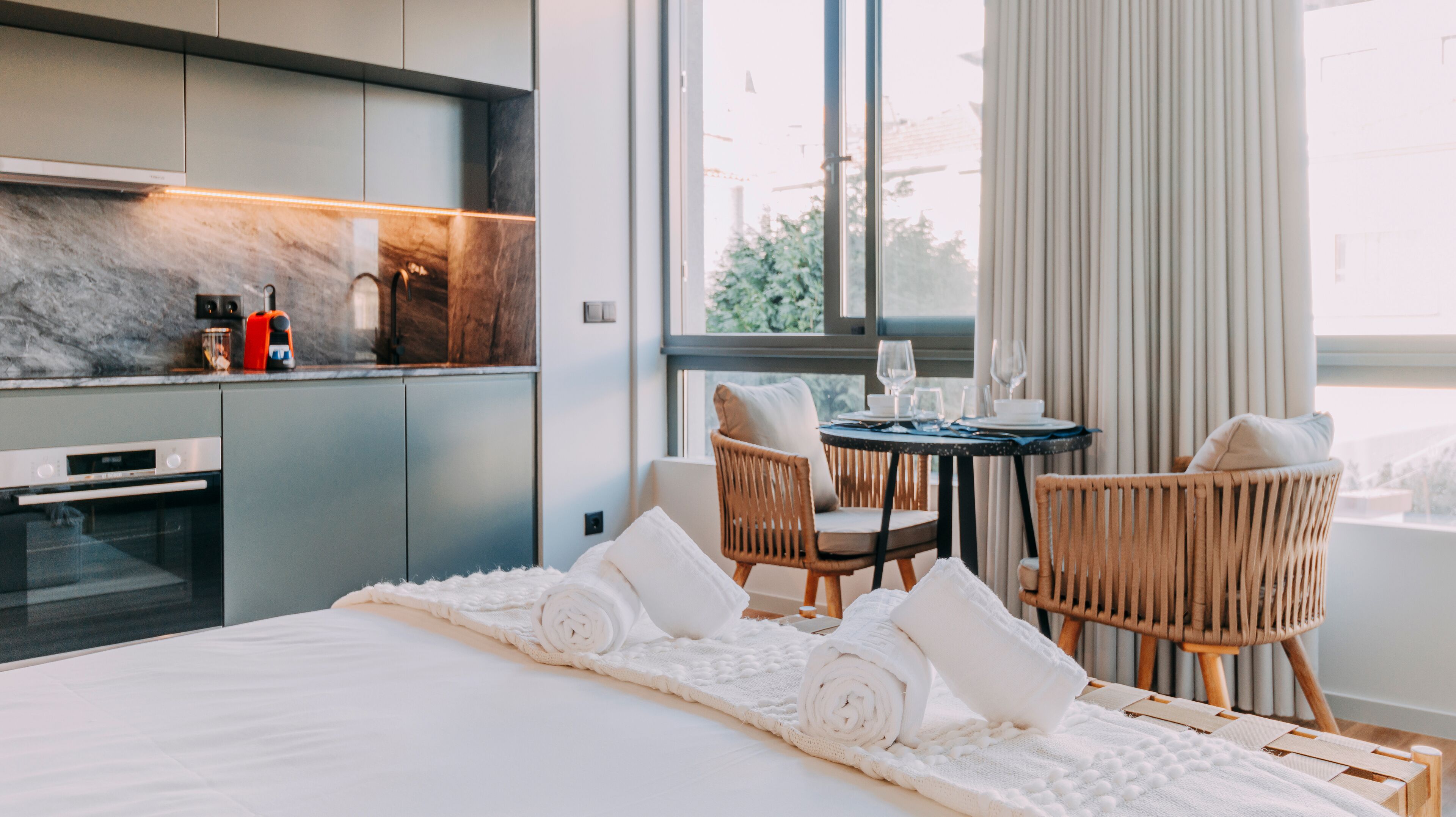 A modern hotel room with a bed, two wicker chairs, a small round table with wine glasses, and a view of the city through large windows.