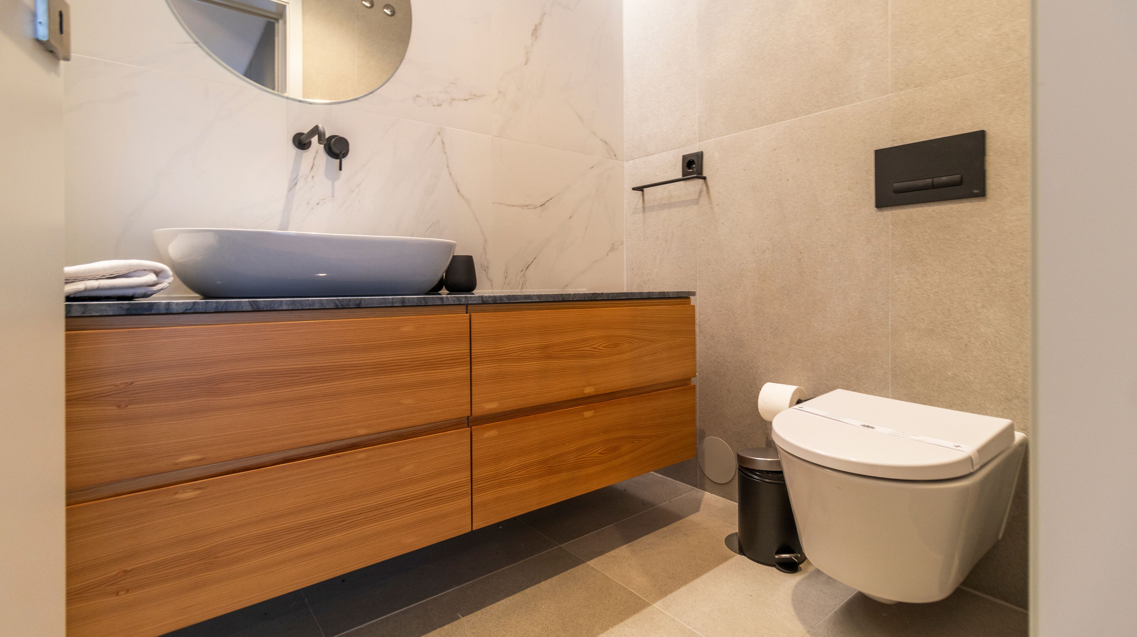 Un baño moderno con tocador de madera, lavamanos sobre encimera, inodoro y cubo de basura.