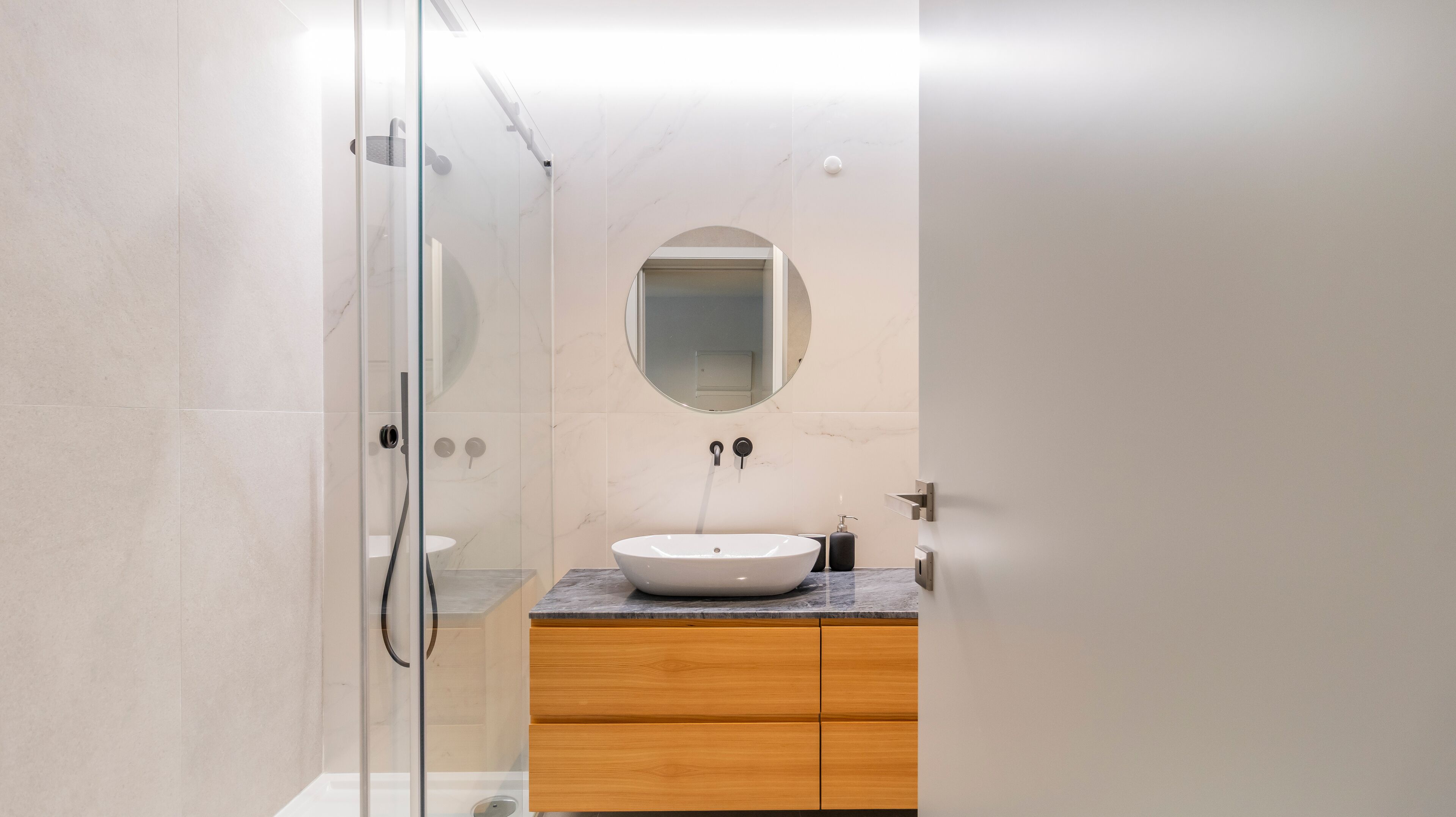 A modern bathroom with a glass-enclosed shower, a round mirror, and a wooden vanity with a white sink.