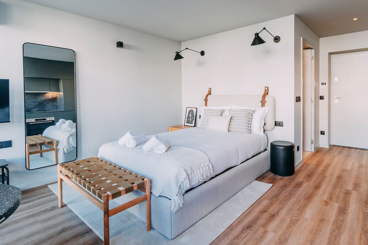 A modern bedroom with a bed, bedside tables, a mirror, a bench, and a trash bin.