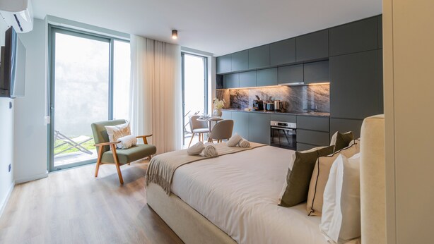 Een moderne hotelkamer met een groot bed, een zithoek met stoelen, een klein keukenhoekje en een balkon met uitzicht.