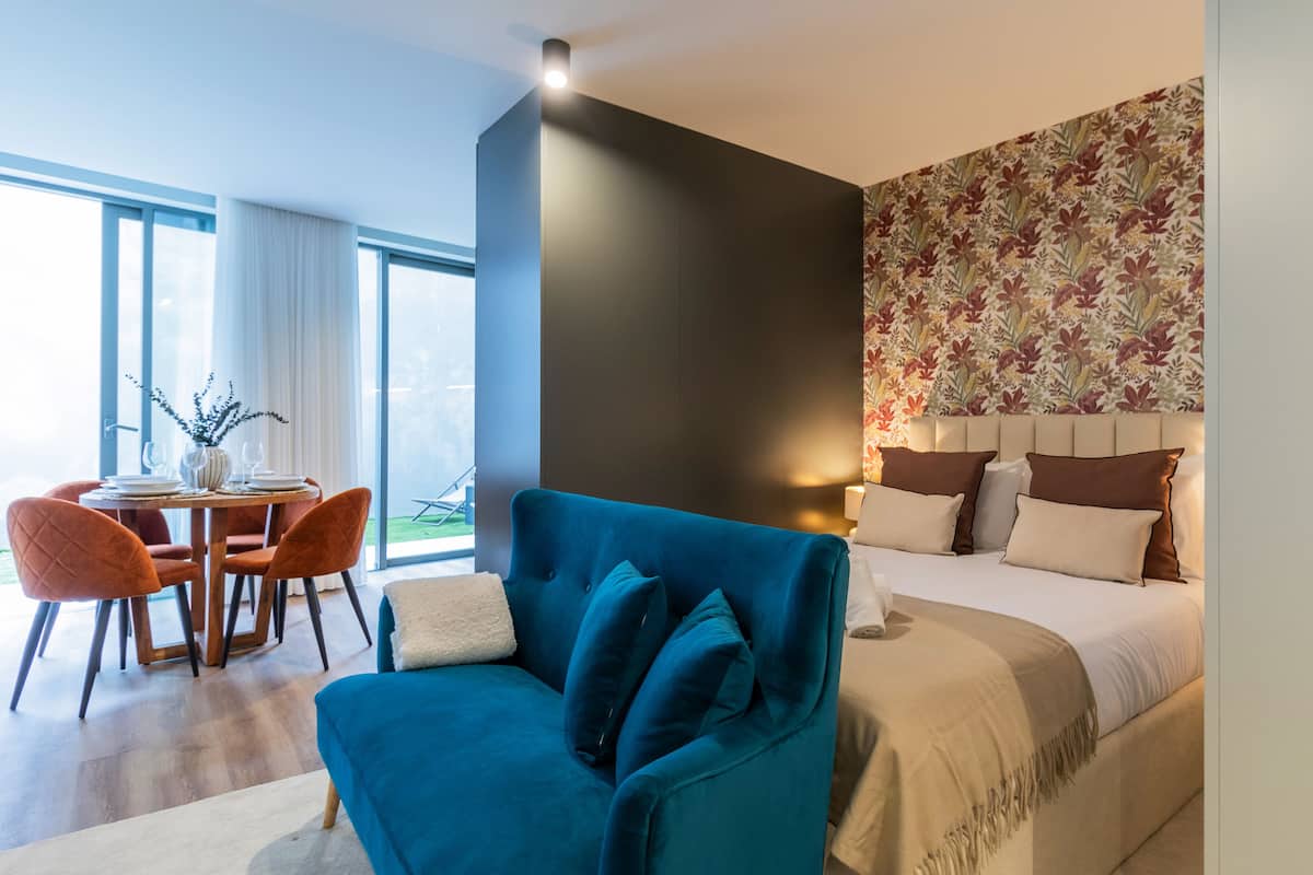 A modern hotel room with a large bed, a blue armchair, a dining table with chairs, and a view of the outdoors.