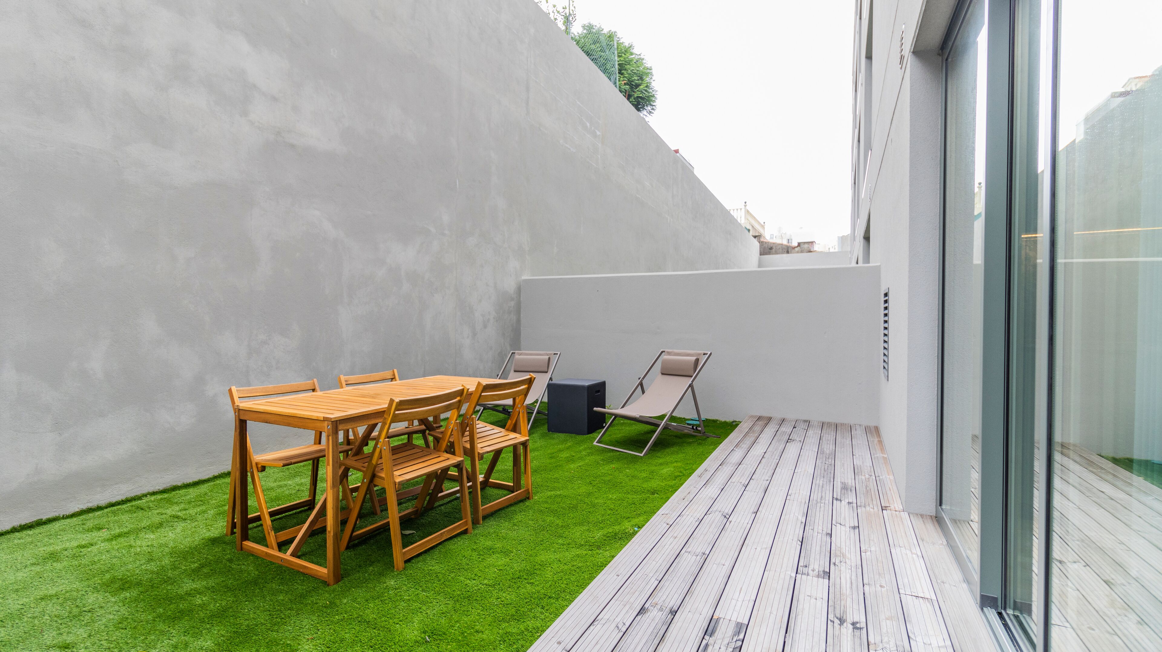 A modern outdoor patio with a wooden table and chairs, a small black speaker, and a grassy area.