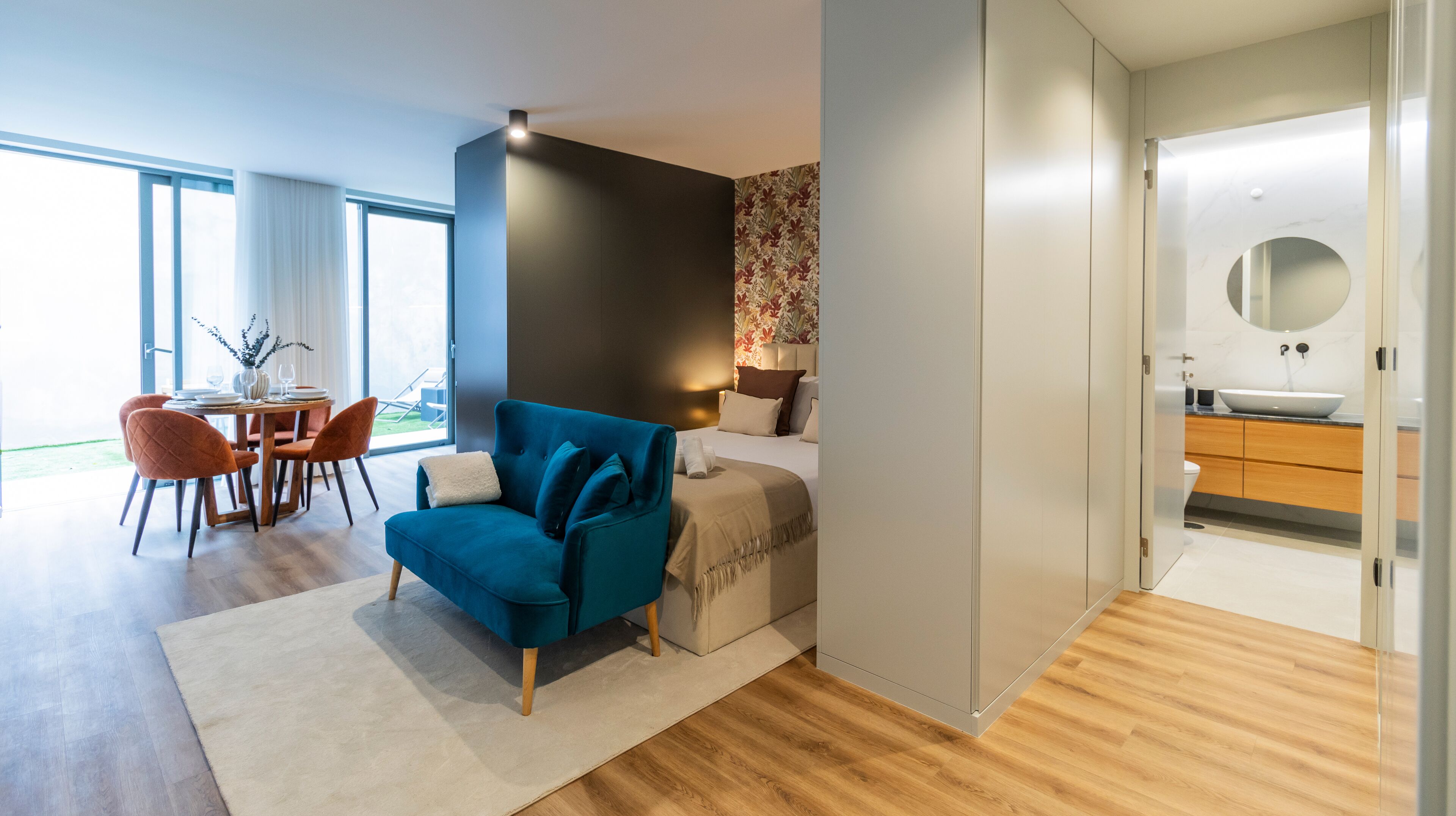 Una habitación de hotel moderna con un sofá azul, una zona de comedor con mesa y sillas, y un baño con un espejo grande y lavamanos.