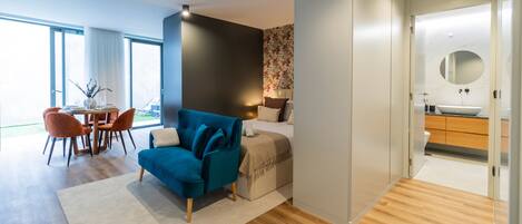Una habitación de hotel moderna con un sofá azul, una zona de comedor con mesa y sillas, y un baño con un espejo grande y lavamanos.
