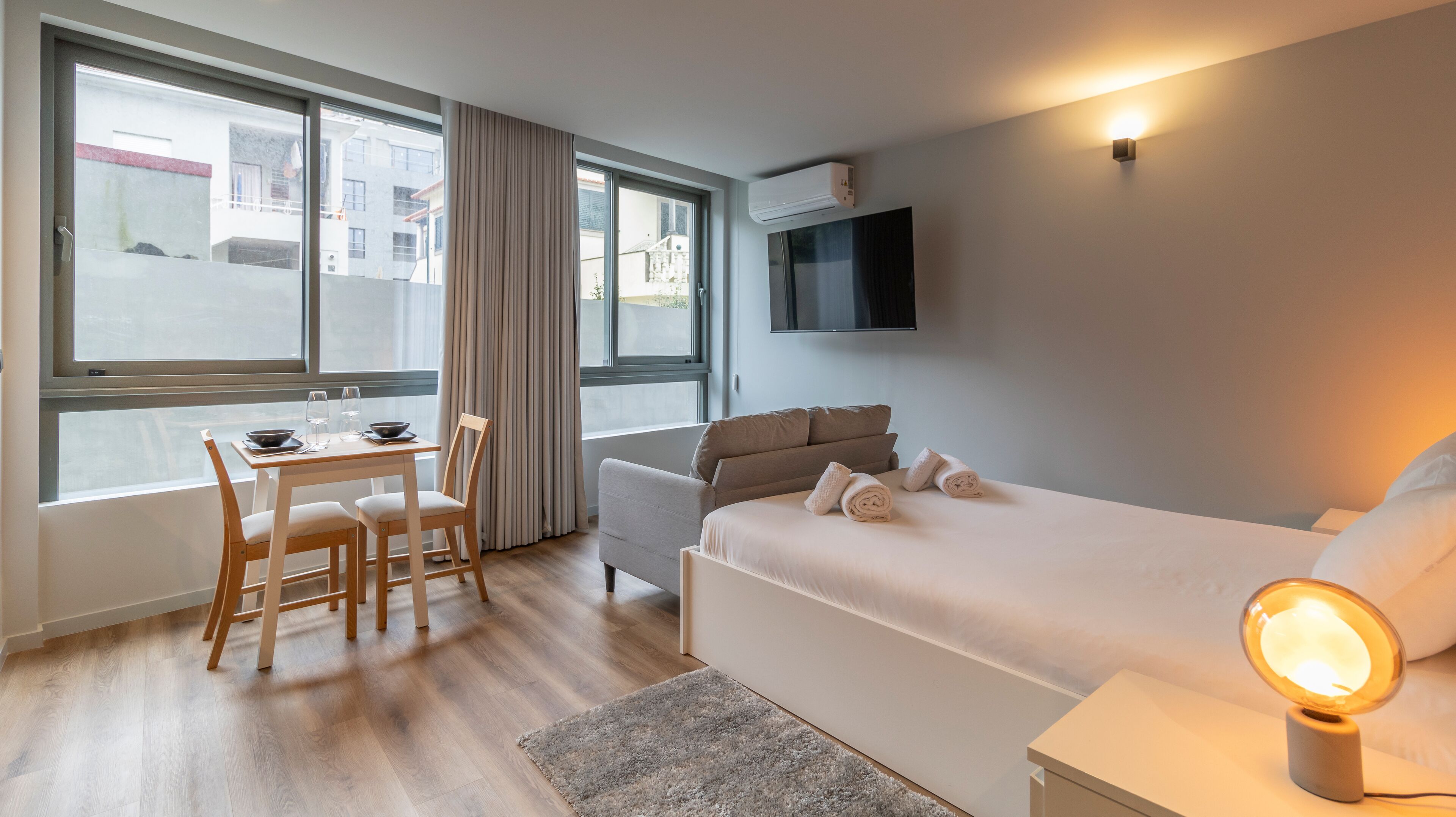 A hotel room with a bed, a desk with chairs, a TV mounted on the wall, and a window with curtains.